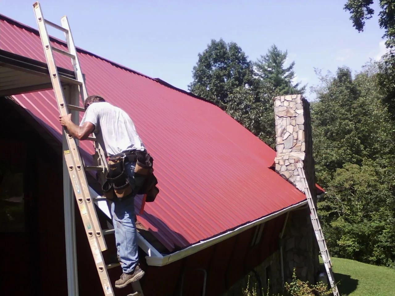 Roofer on a ladder beside a red metal roof and stone chimney — Soffit & Fascia Repair by Roofing Brothers in Beckley